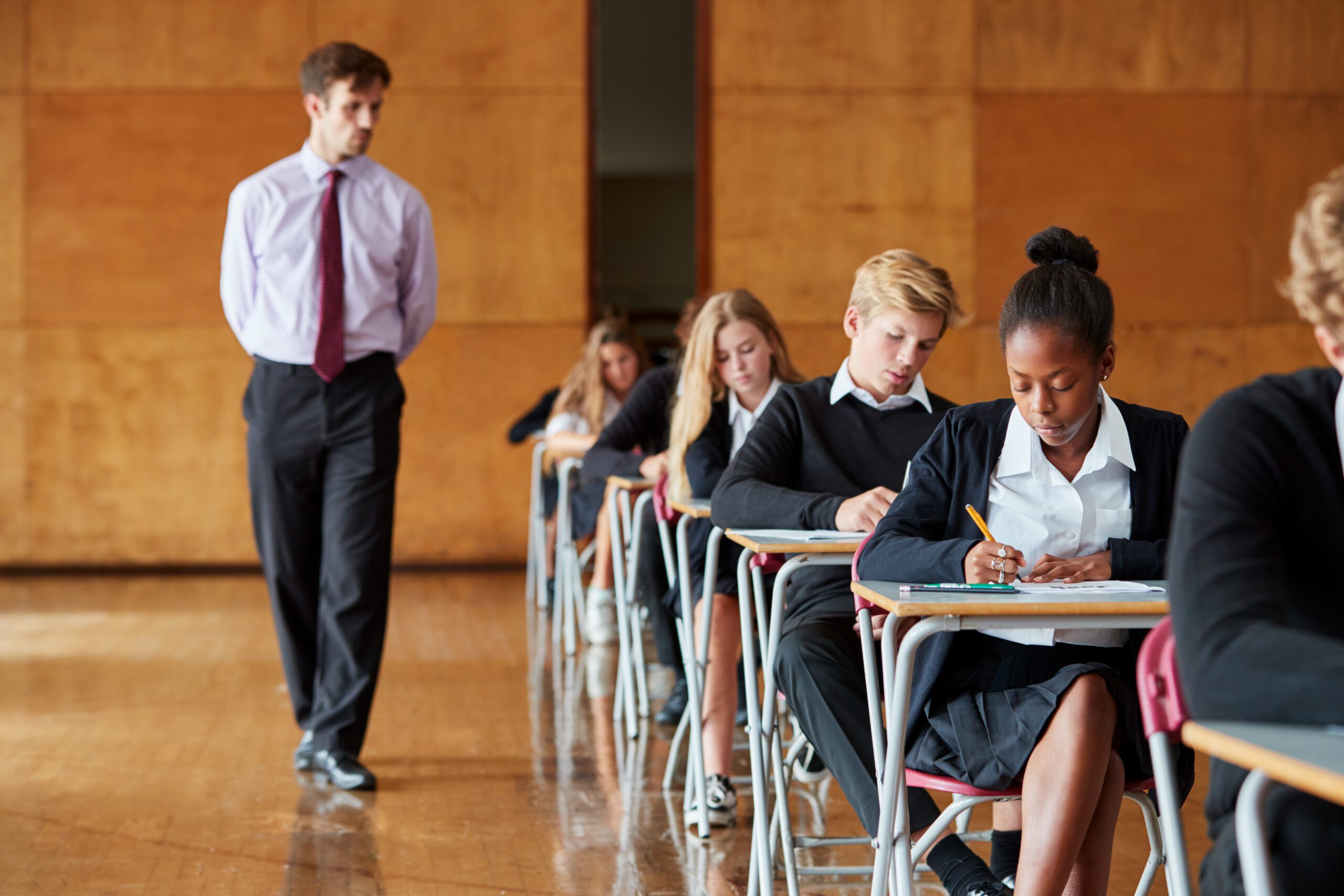 Header image for exam ready handwriting with man walking down a line of desks whilst students are writing at exam tables.