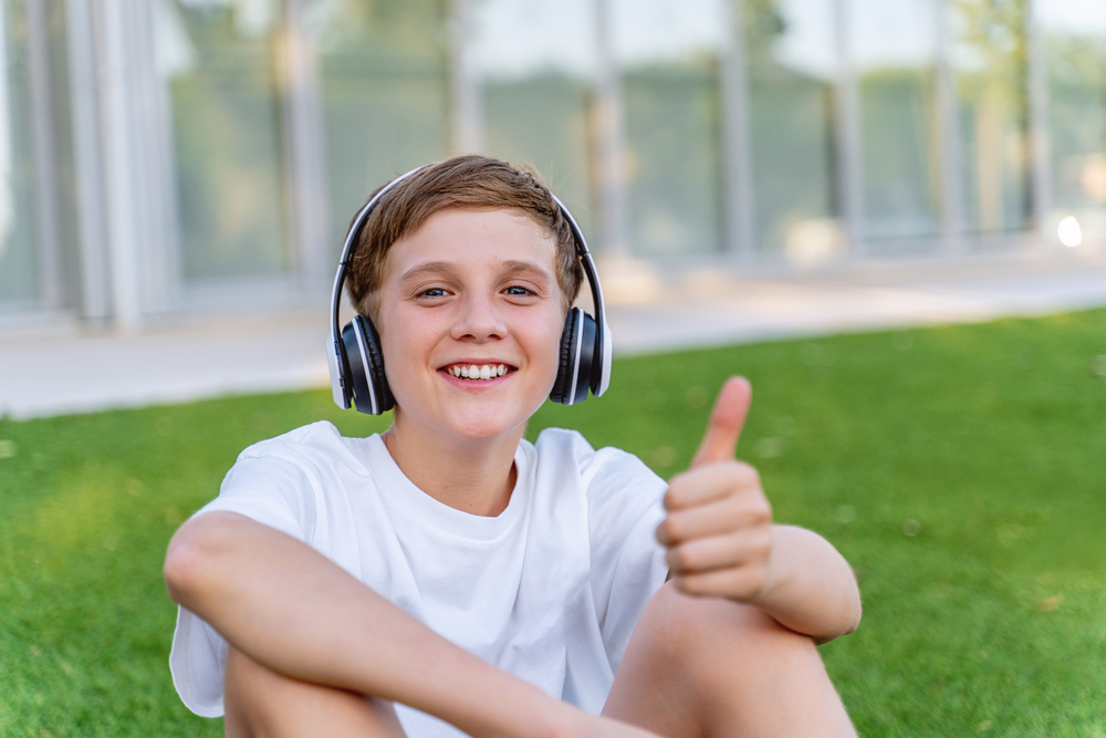 Young boy with headphones showing thumb up and smiling while sitting outdoors used on readable handwriting page.